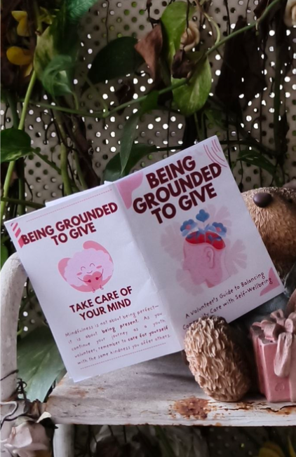 Physical printed copies of Being Grounded to Give minibook displayed on a shelf with plants and a teddy bear